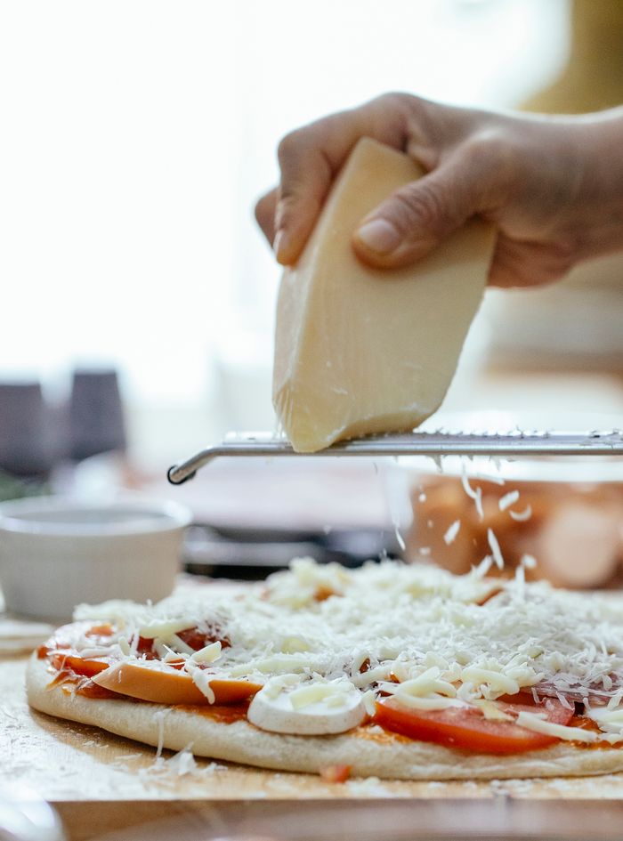 Unrecognizable cook grating cheese on raw pizza with toppings placed on table during cooking process in kitchen against blurred background
